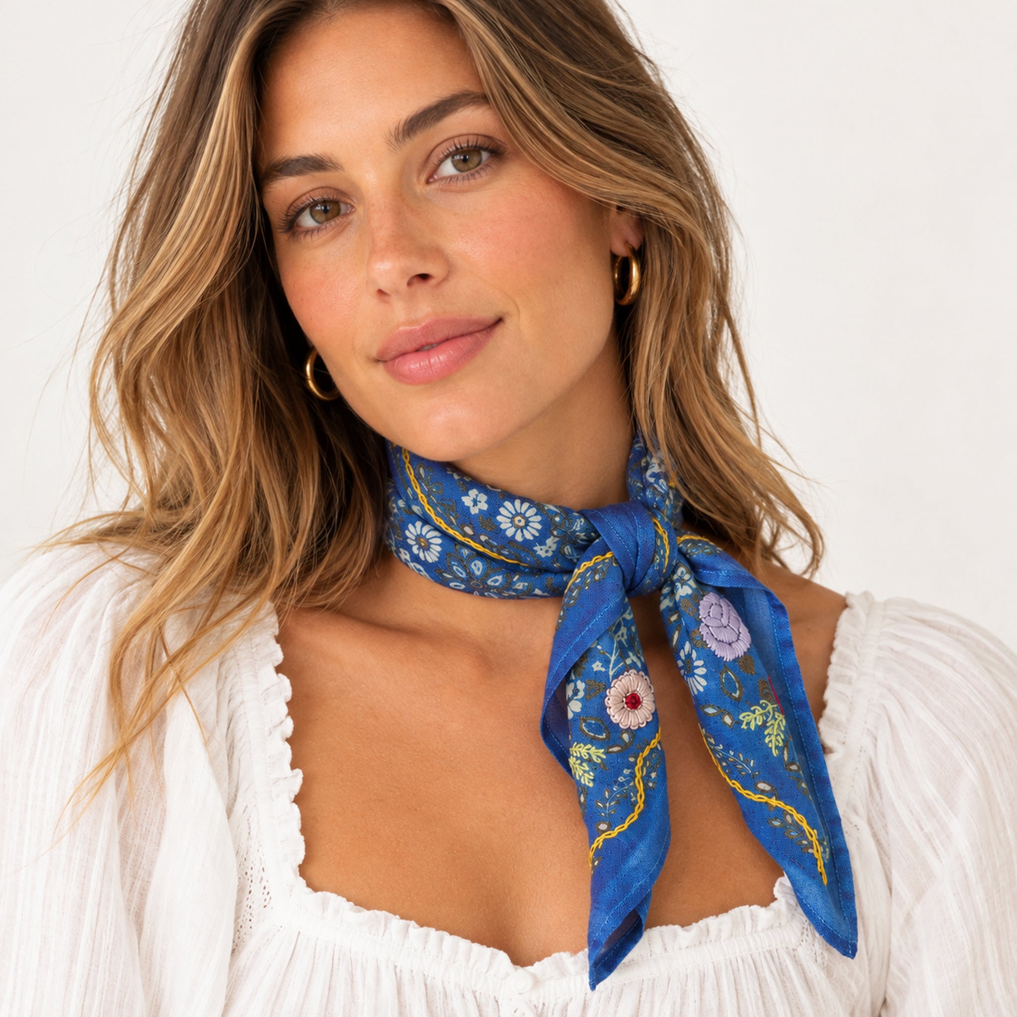 A woman wearing a blue patterned silk scarf tied around her neck, paired with a white ruffled blouse and gold hoop earrings, against a clean white background.