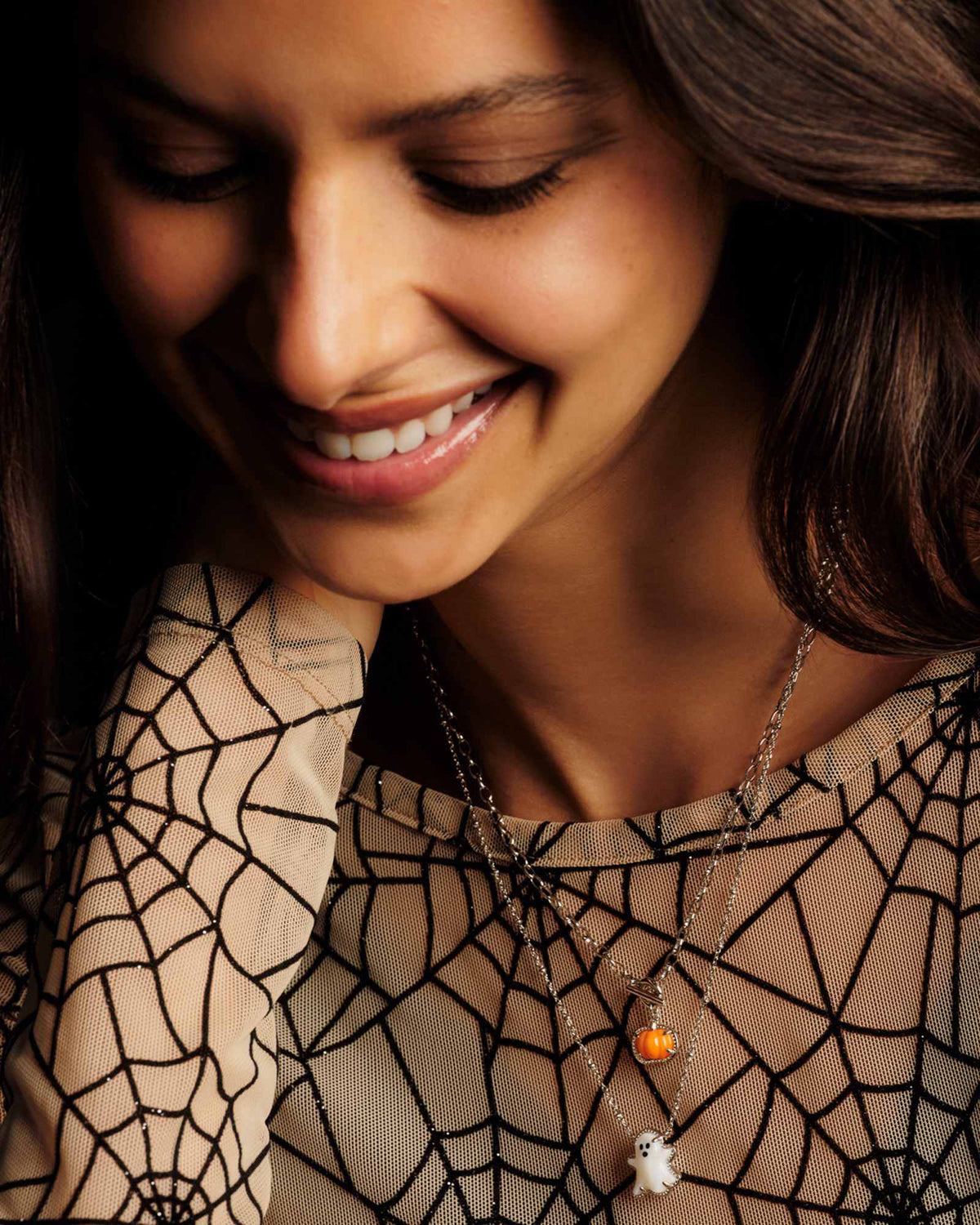 A close-up lifestyle shot of a woman wearing a double-layered silver necklace with a small orange pumpkin and a white ghost pendant, styled with a black spiderweb-patterned top.