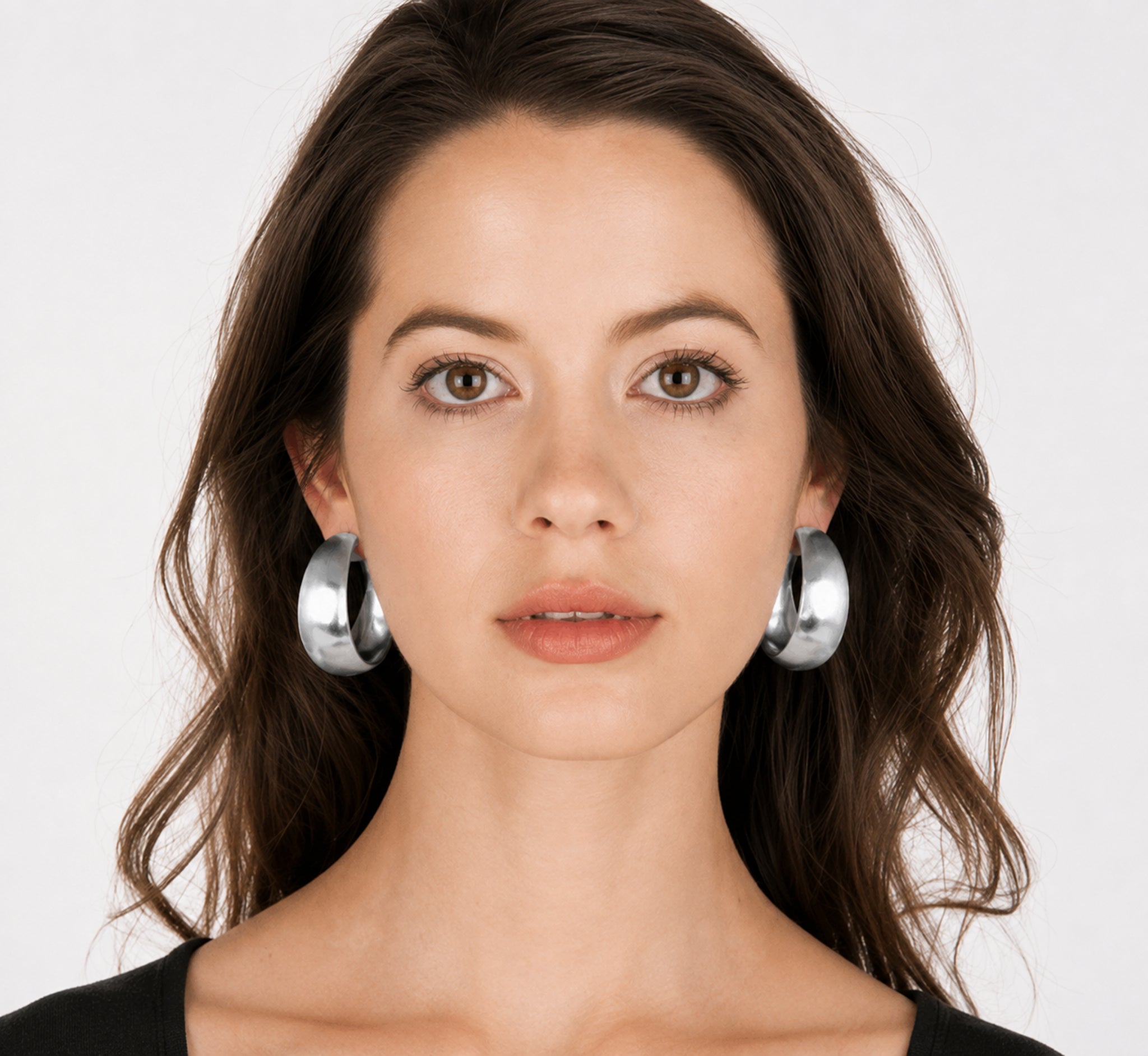 A close-up portrait of a woman with dark brown hair wearing large, polished silver hoop earrings against a clean white background.