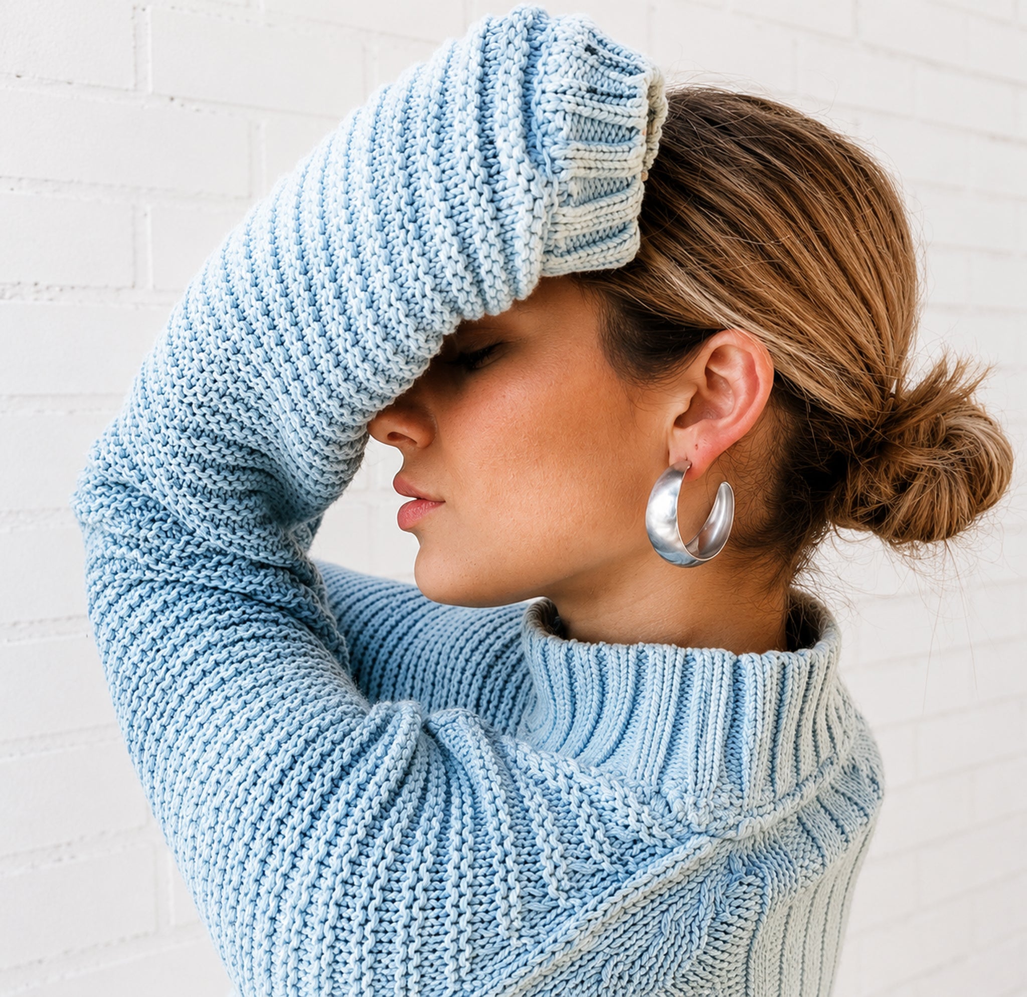 A woman wearing a light blue chunky knit turtleneck sweater and large silver hoop earrings, posing with her arm raised against a white brick wall.
